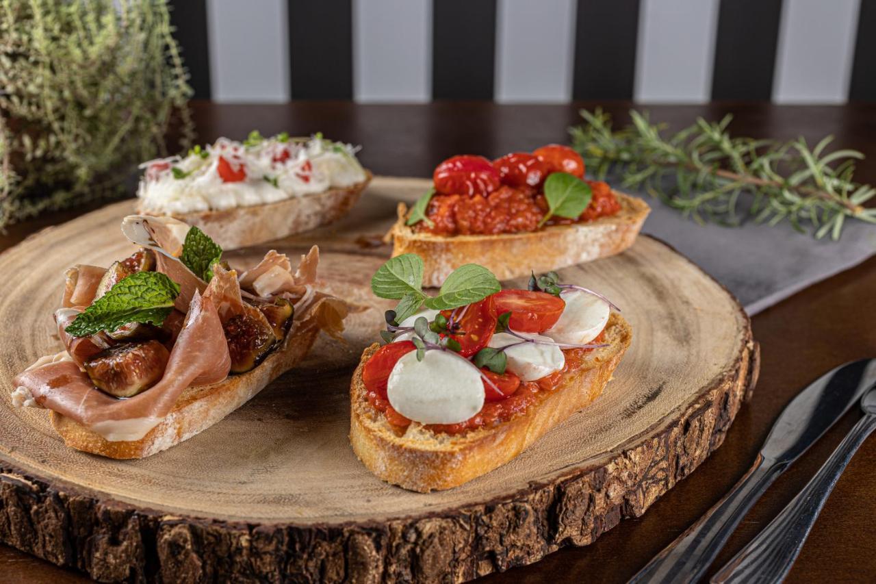 Buona Vitta Gramado Resort & Spa By Gramado Parks Bagian luar foto The photo displays a wooden serving platter featuring four different types of bruschetta. Each piece of bruschetta is topped with various ingredients:

1. The top left has a creamy spread, possibly cheese or ricotta, garnished with fresh herbs and sm