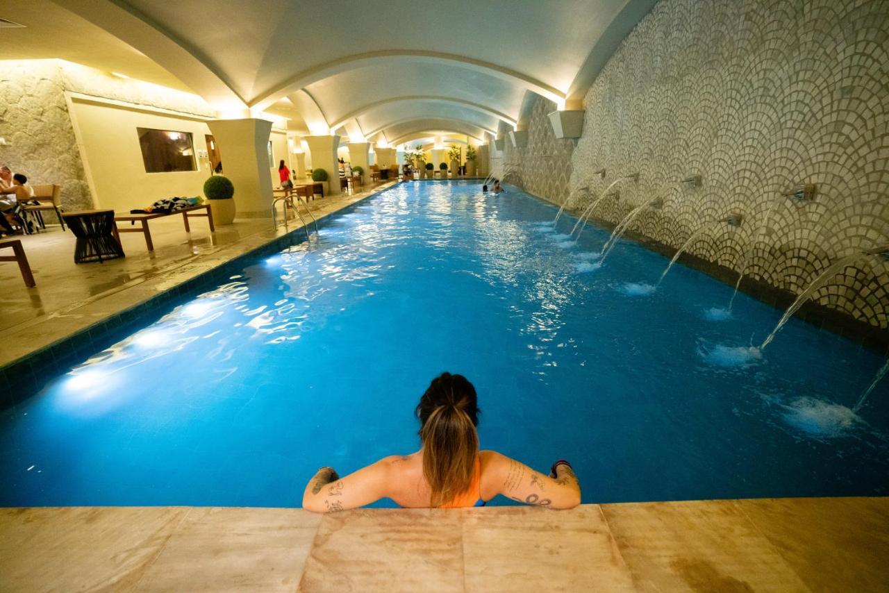 Buona Vitta Gramado Resort & Spa By Gramado Parks Bagian luar foto The photo shows an indoor swimming pool with a long, narrow design. The water is a clear blue, and there are features such as fountains along one side. In the foreground, a person with long hair is sitting on the edge of the pool, looking out over th
