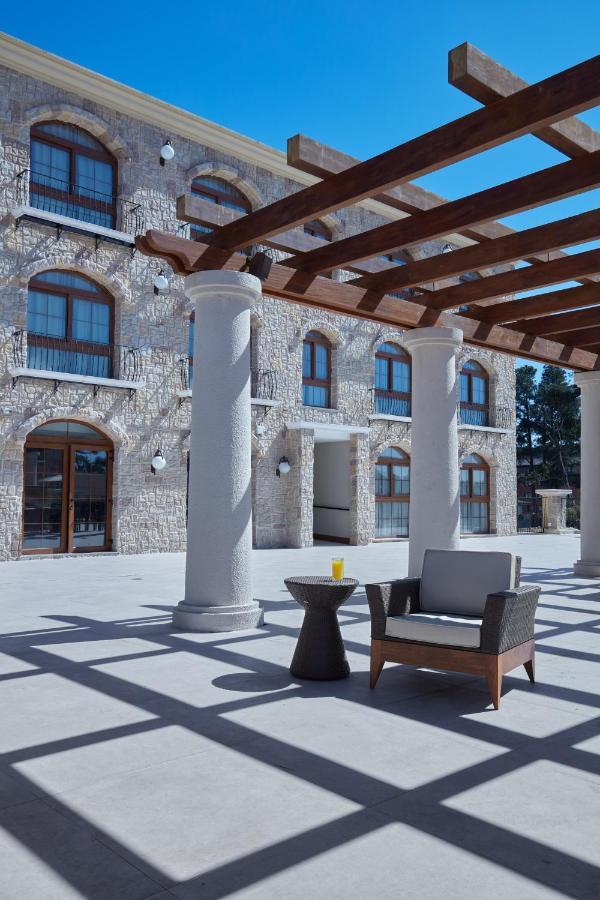 Buona Vitta Gramado Resort & Spa By Gramado Parks Bagian luar foto The photo shows a well-designed outdoor space with a stone wall building in the background. There are several tall columns that support a wooden pergola overhead, creating a shaded area. In this space, there is a stylish chair and a small table besid
