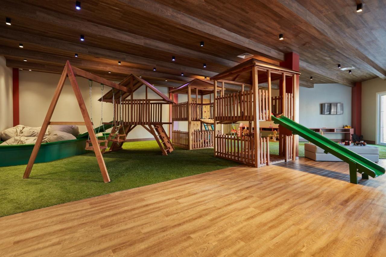 Buona Vitta Gramado Resort & Spa By Gramado Parks Bagian luar foto The photo shows an indoor play area designed for children. It features a wooden play structure that includes multiple levels, slides, and a swing set. The floor is covered with wooden planks, and there is a green carpet-like area that adds a soft tou