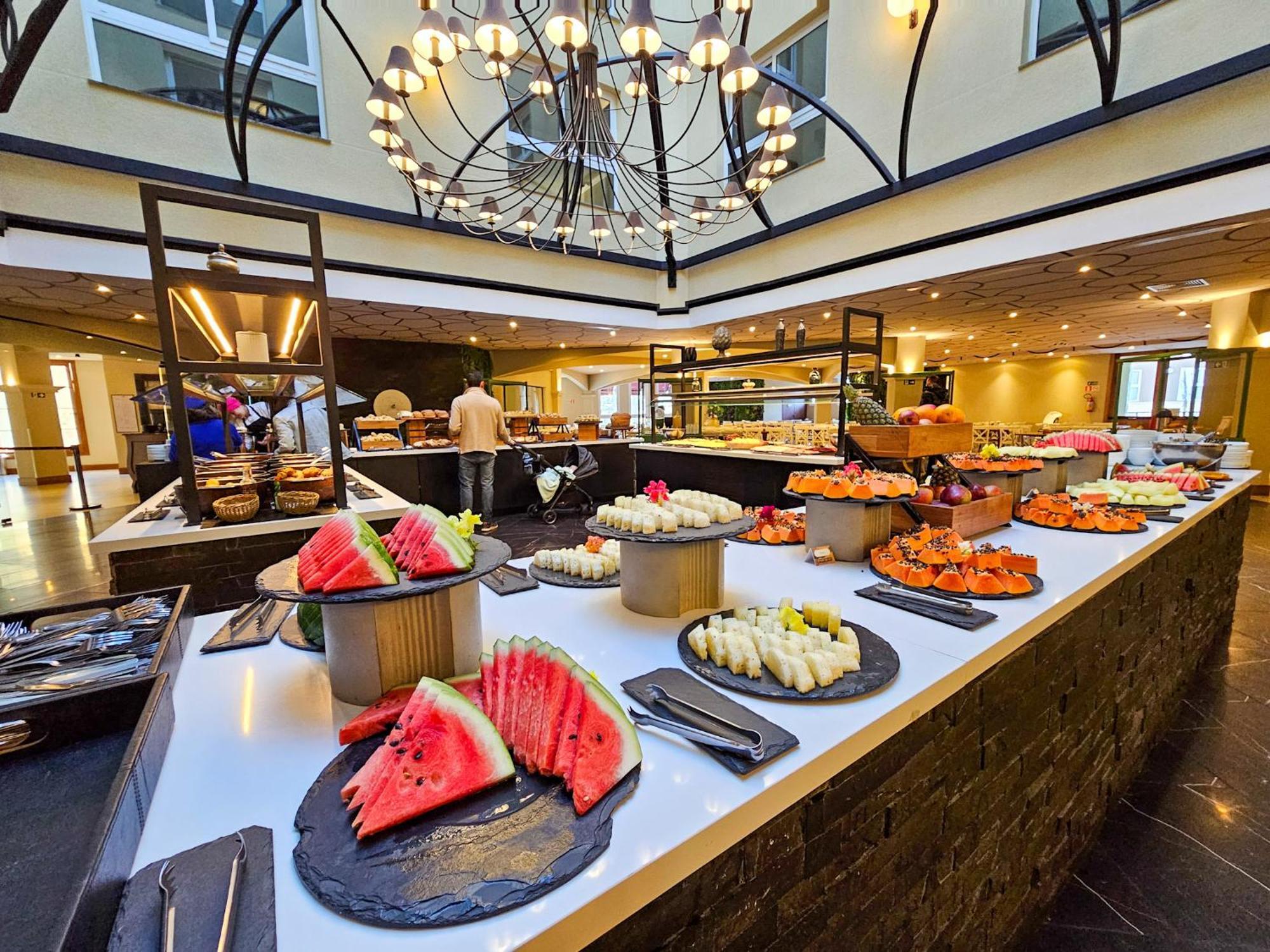Buona Vitta Gramado Resort & Spa By Gramado Parks Bagian luar foto The photo depicts a lavish buffet setup in a dining area. In the foreground, there are several platters featuring an assortment of colorful food items, primarily fruits like watermelon and various types of sushi or rolls. The buffet table is elegantl