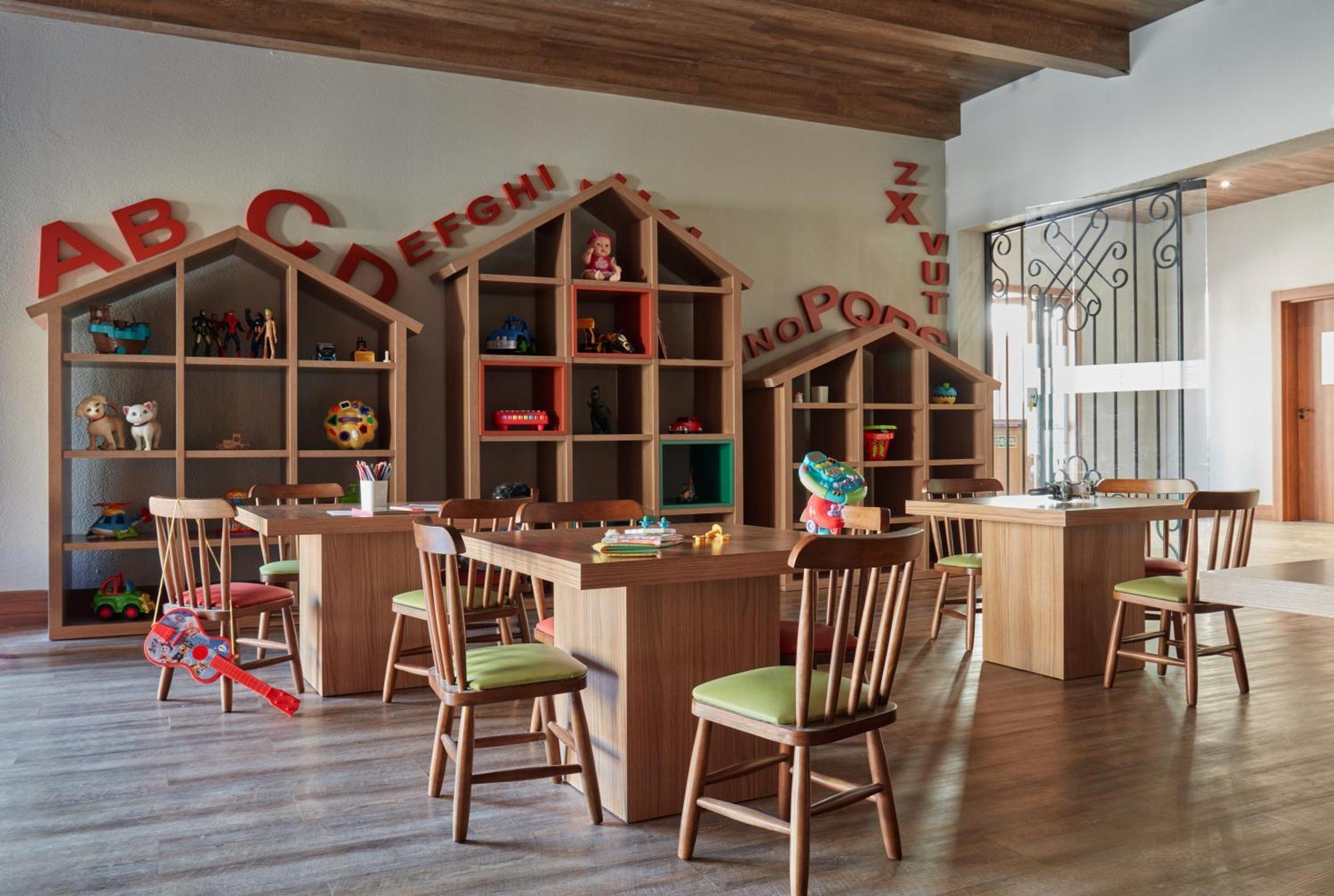 Buona Vitta Gramado Resort & Spa By Gramado Parks Bagian luar foto The photo shows a classroom or playroom designed for children. It features wooden furniture, including several tables and chairs with green cushions. In the background, there are large, open shelving units shaped like houses, displaying various toys 