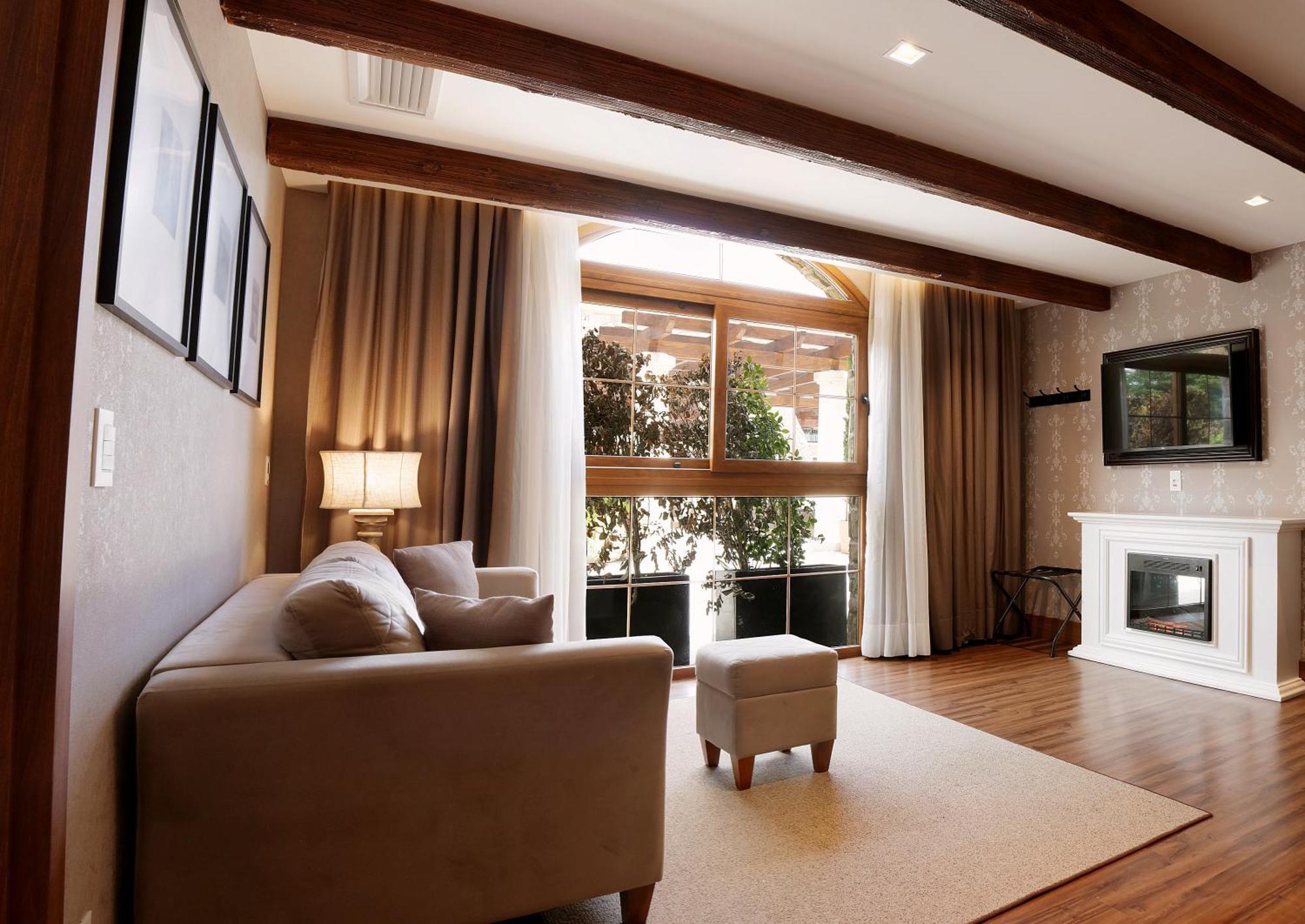 Buona Vitta Gramado Resort & Spa By Gramado Parks Bagian luar foto The photo depicts a cozy living room setting. It features a light-colored couch, a small ottoman, and a floor lamp beside the couch. Large windows are covered with curtains, allowing natural light to illuminate the space. In the background, there is 