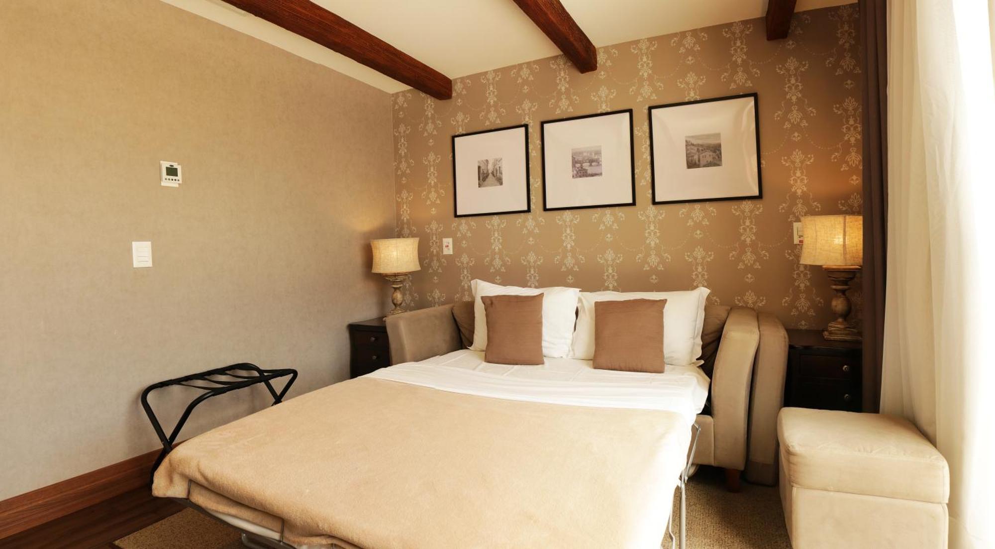 Buona Vitta Gramado Resort & Spa By Gramado Parks Bagian luar foto The photo shows a cozy bedroom with a neutral color palette. There is a bed with white linens and two brown decorative pillows. On either side of the bed, there are bedside tables with lamps. The walls feature a subtle floral pattern, and three frame