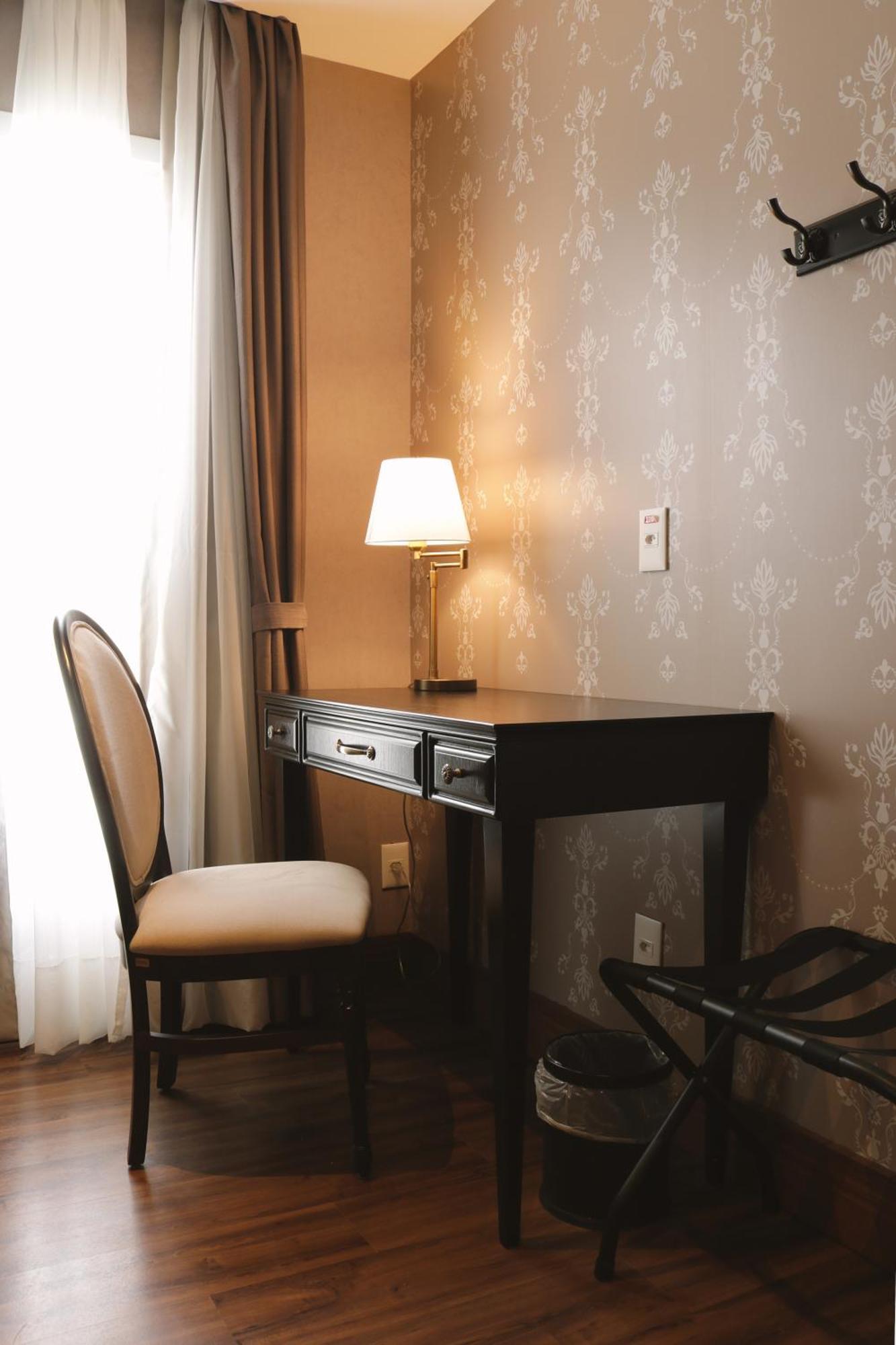 Buona Vitta Gramado Resort & Spa By Gramado Parks Bagian luar foto The photo shows a small desk set against a wall with a decorative pattern. The desk is black with a few drawers and has a small lamp on it. Next to the desk, there is a chair with a light-colored cushion. Soft light is coming through a window, castin