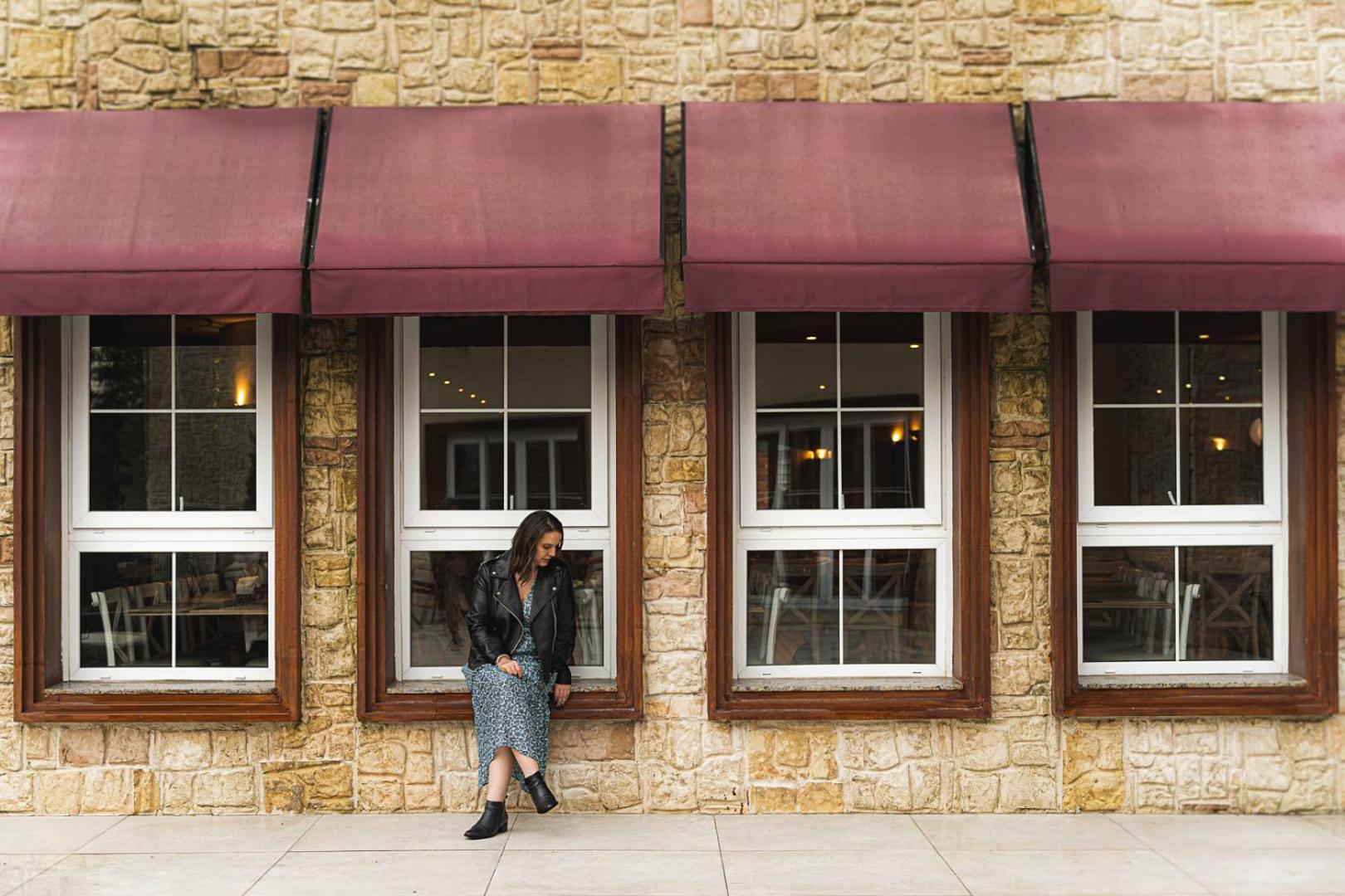 Buona Vitta Gramado Resort & Spa By Gramado Parks Bagian luar foto The photo shows a woman standing against a stone wall with three large windows in front of her, each framed with wooden borders. The windows have maroon awnings above them. The woman is dressed in a long skirt and leather jacket, leaning slightly aga